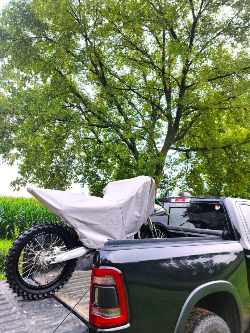Dirt bike covered with a protective tarp in a truck bed under a large tree.