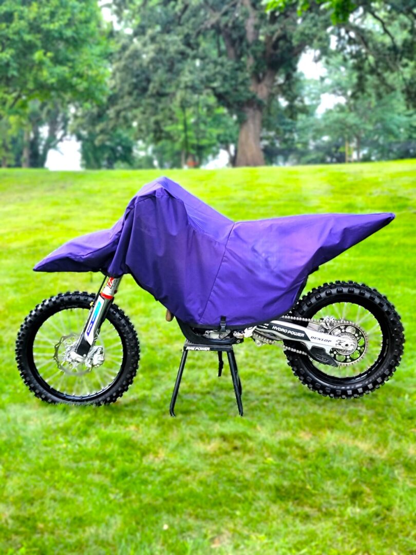 A dirt bike covered with a purple protective cover in a grassy area.